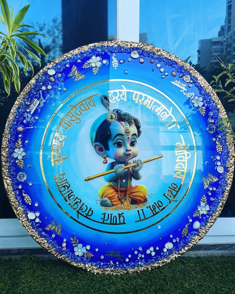 A round blue resin wall plate with gold lettering, a small Krishna figure, shells, pearls and a gold textured rim